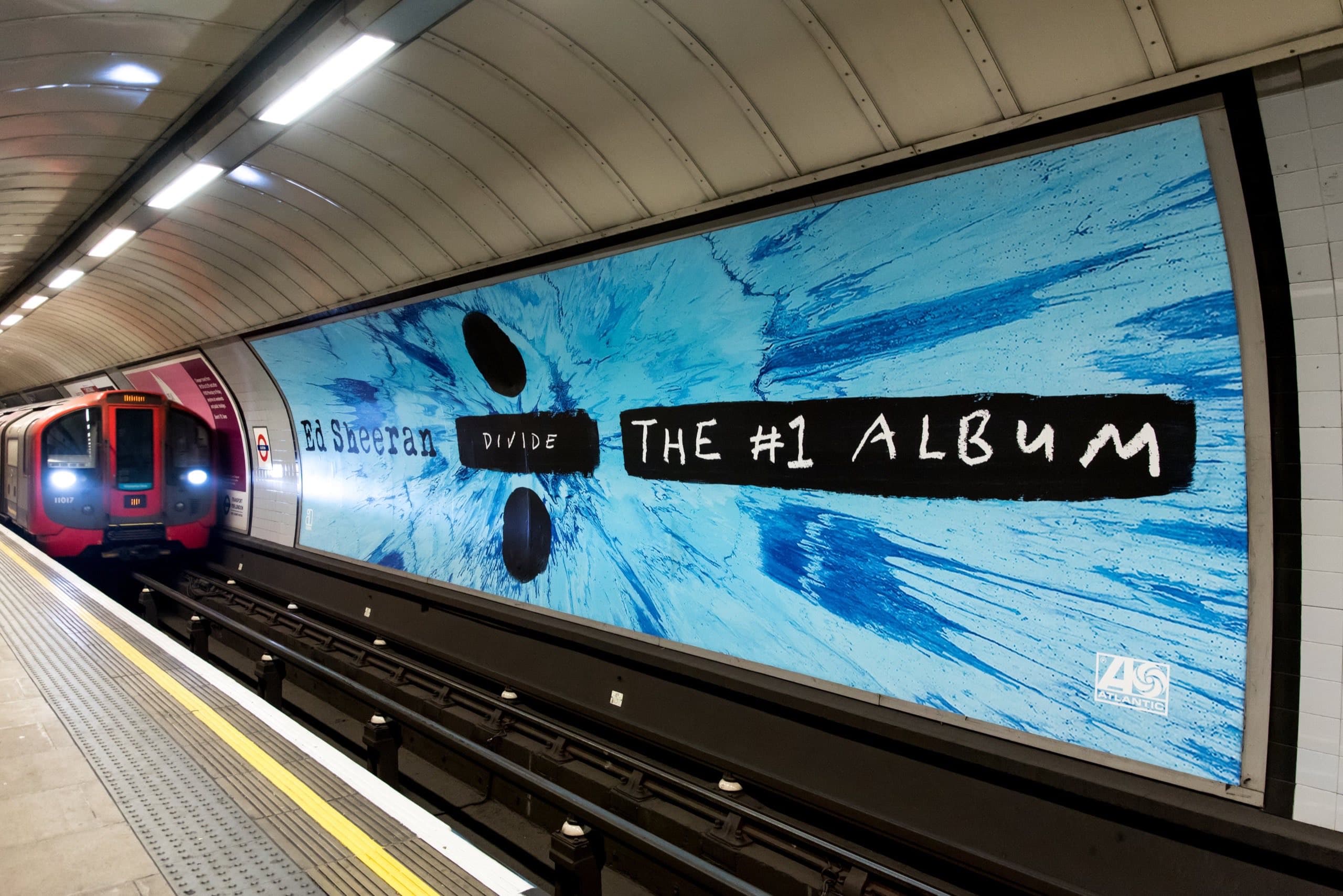 A cross-track 96-sheet poster running the length of a London Underground platform wall, carrying an Ed Sheeran album advertisement, with a train arriving alongside it.