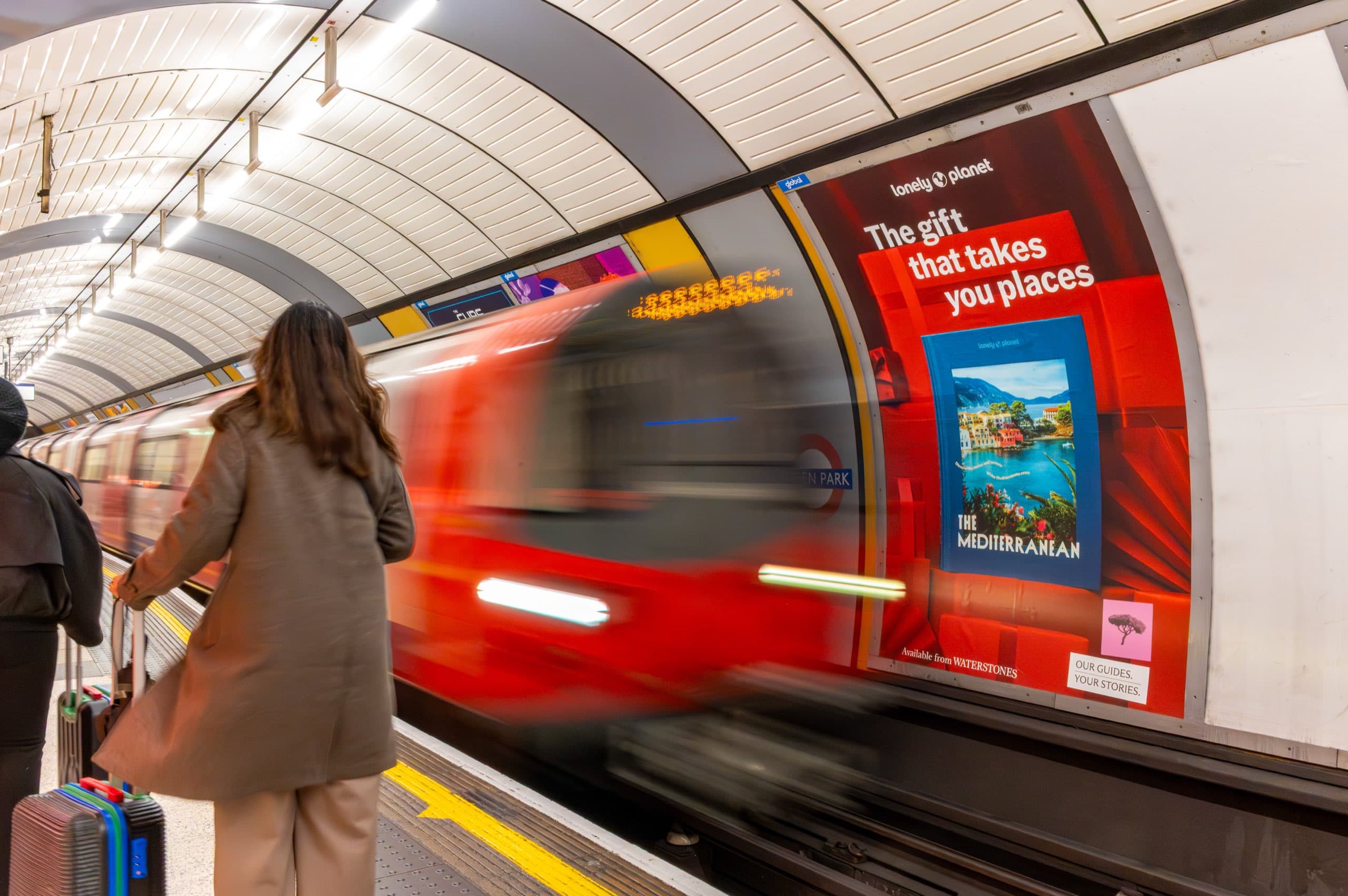 A portrait Lonely Planet poster reading “The gift that takes you places” on the cross-track wall at Green Park, with a train passing and passengers waiting on the platform.