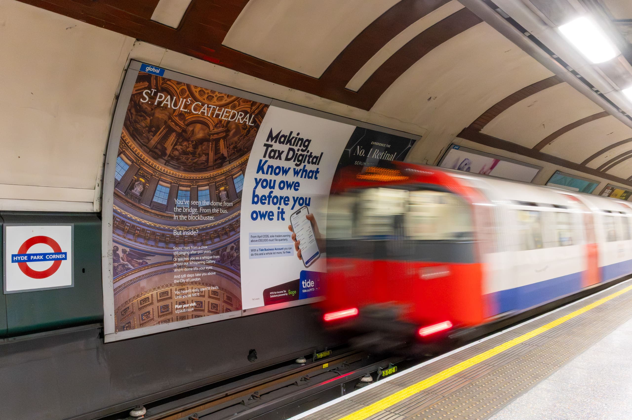 A cross-track poster site at Hyde Park Corner carrying a St Paul's Cathedral advertisement, with a Piccadilly line train passing the platform.