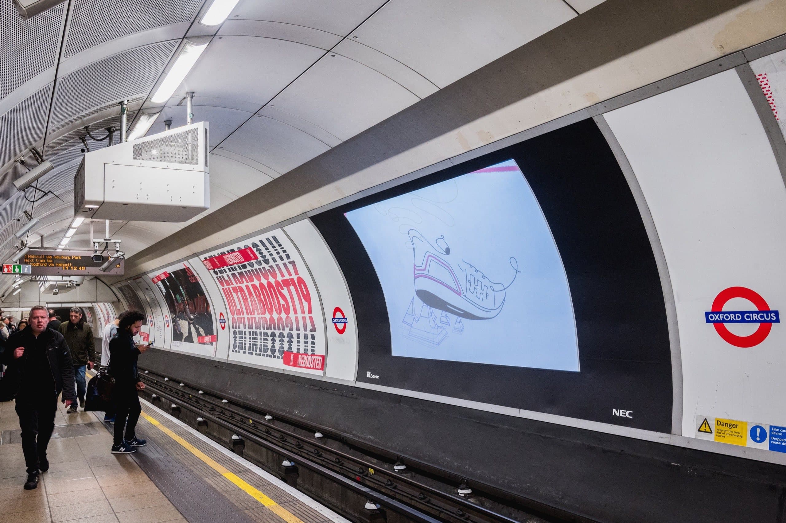 A digital 48-sheet screen carrying an Adidas advertisement on the trackside wall at Oxford Circus, with printed cross-track posters running alongside it and passengers waiting on the platform opposite.