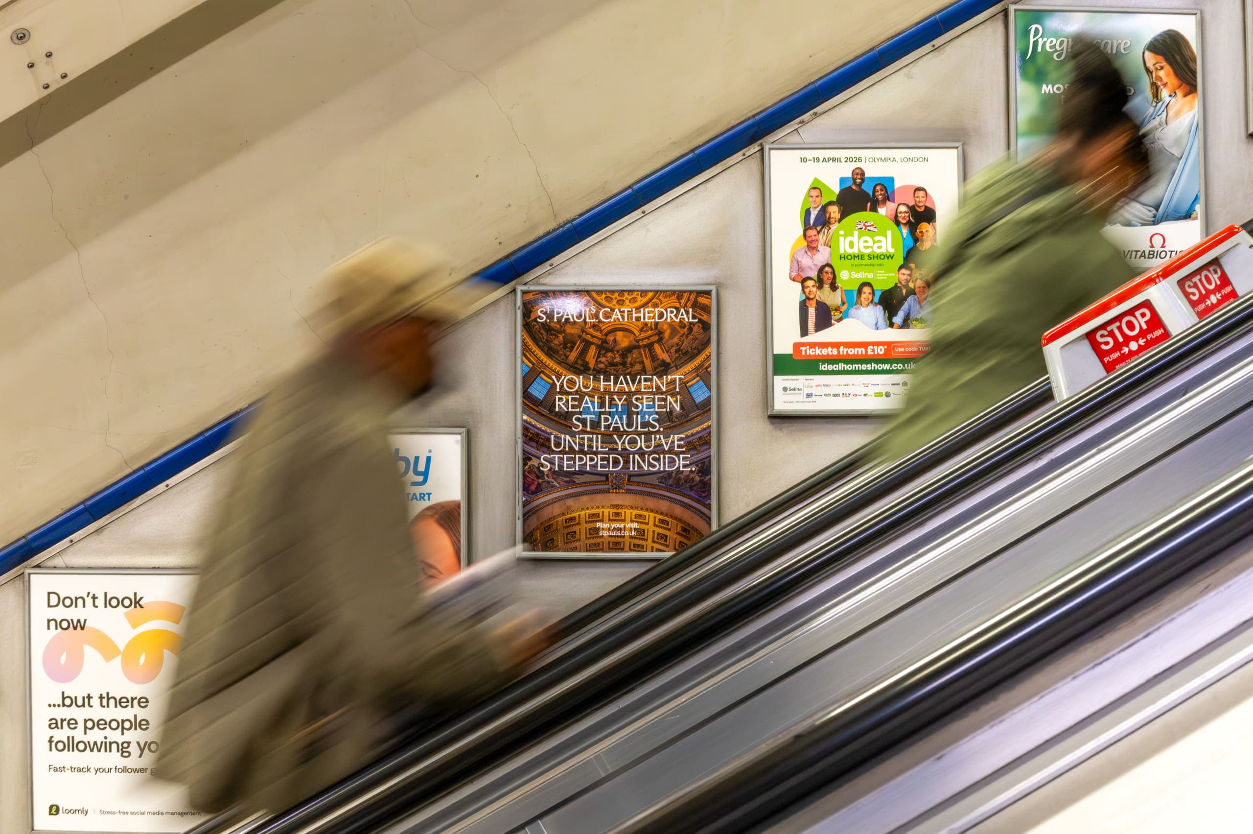 Static escalator panels beside an Underground escalator, the central one carrying a St Paul's Cathedral advertisement, as a passenger rides past.