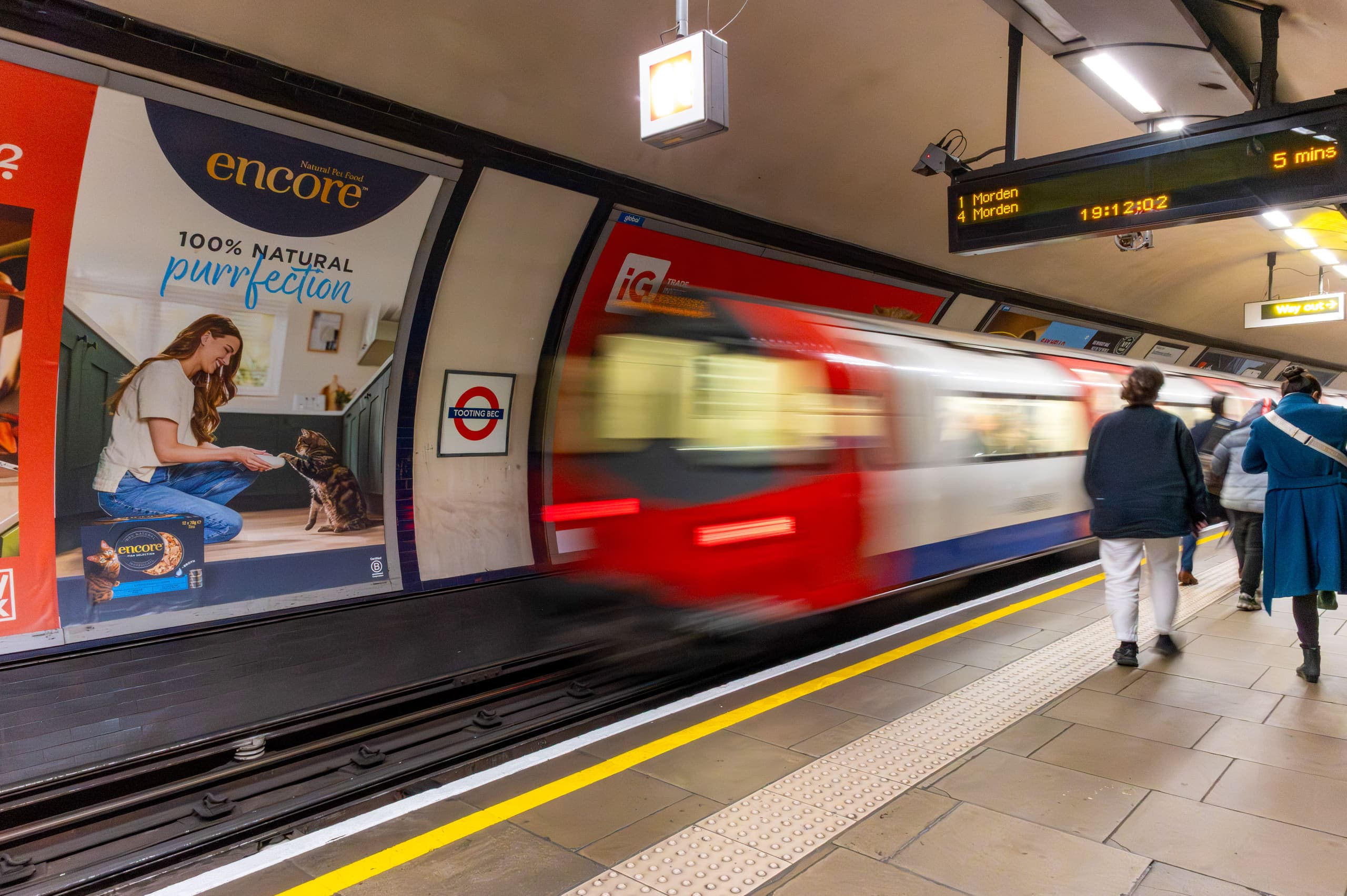 A cross-track 16-sheet poster advertising Encore on the platform wall at Tooting Bec, with a southbound Northern line train leaving.