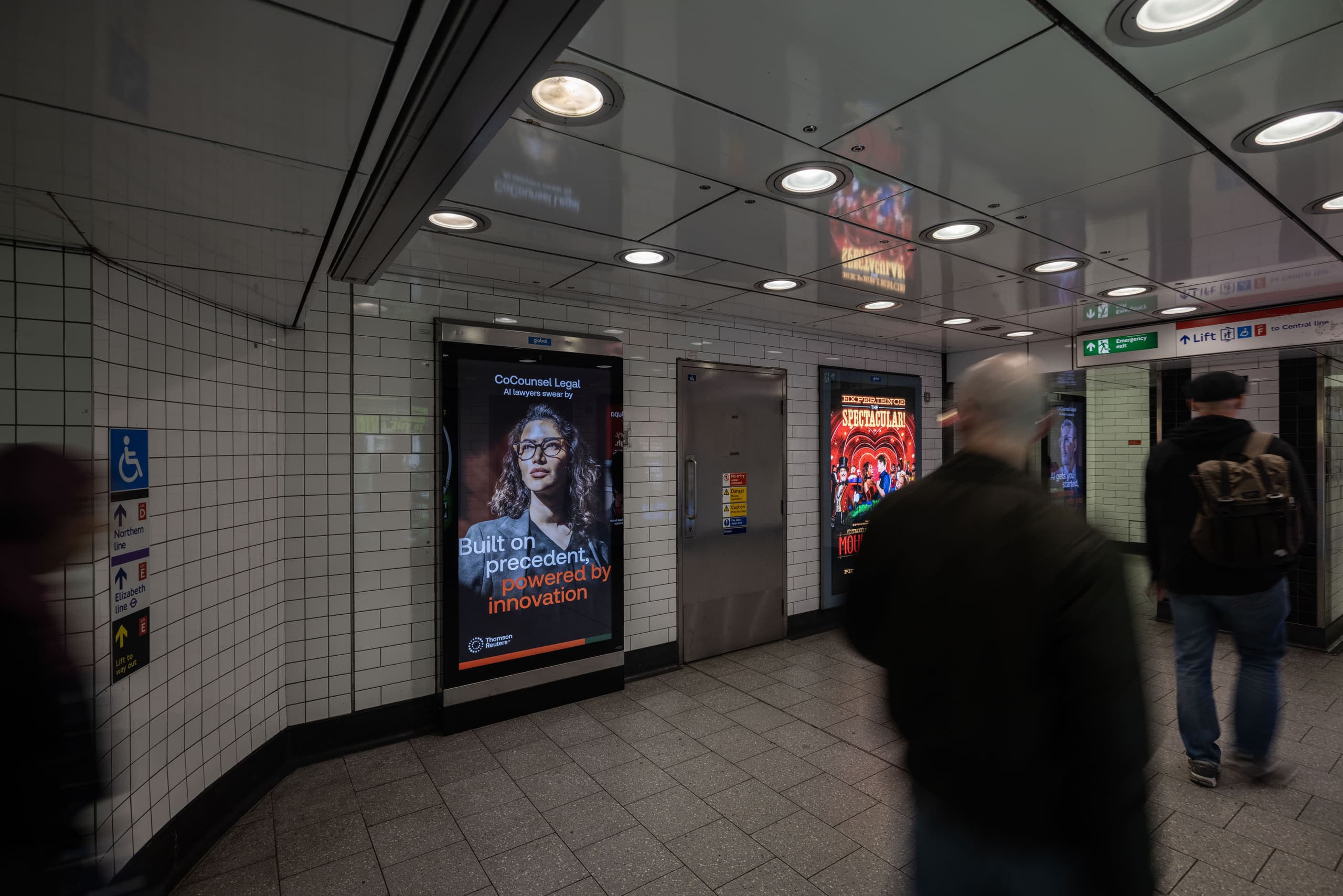 A portrait digital 6-sheet screen carrying a Thomson Reuters advertisement in a tiled corridor at Tottenham Court Road, beside wayfinding for the Northern, Central and Elizabeth lines.