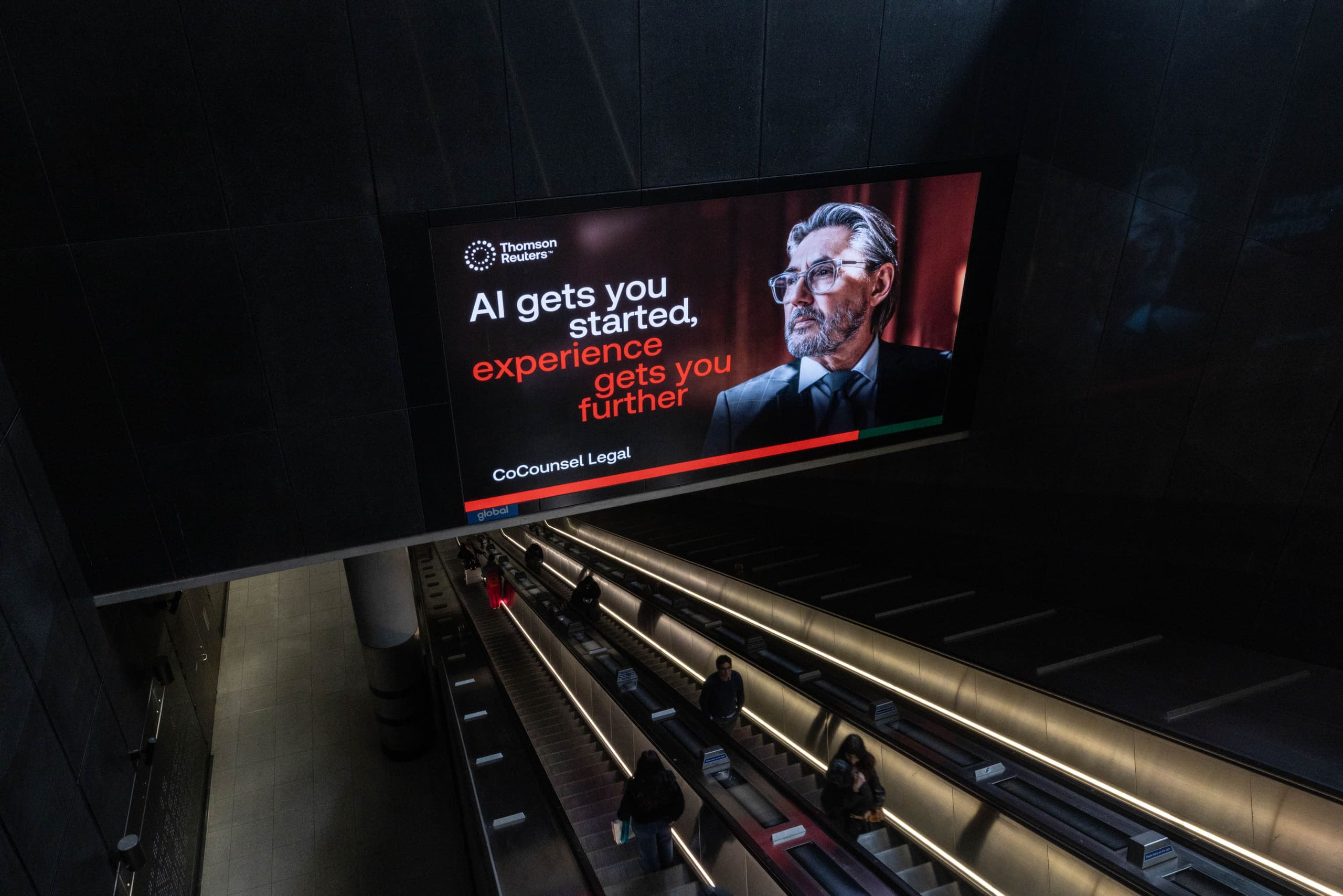 A digital screen above an Underground escalator hall carrying a Thomson Reuters advertisement, with passengers riding down towards the platforms.