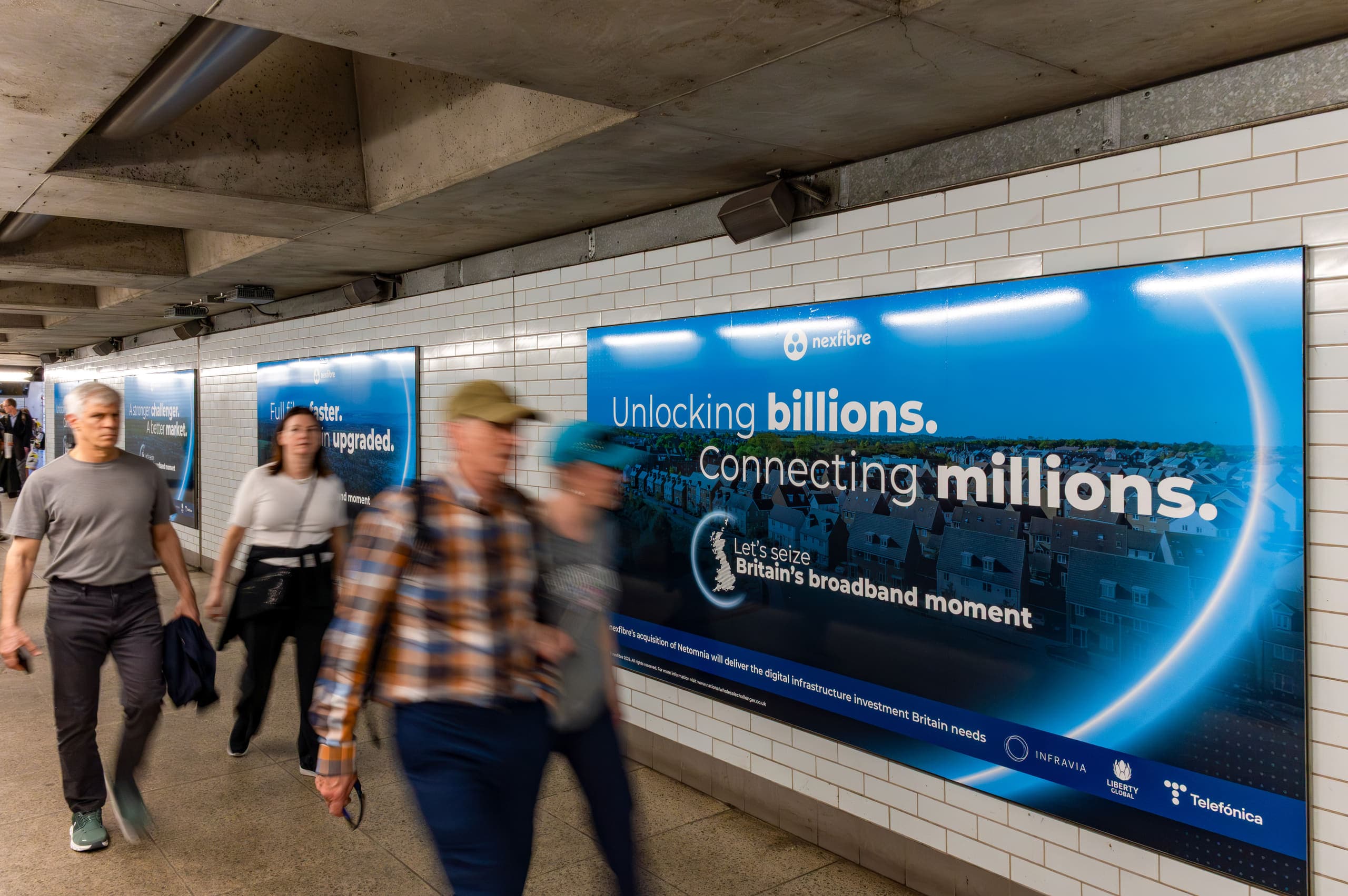 A large nexfibre panel on the tiled wall of an Underground walkway with its creative fully legible, as commuters walk past.