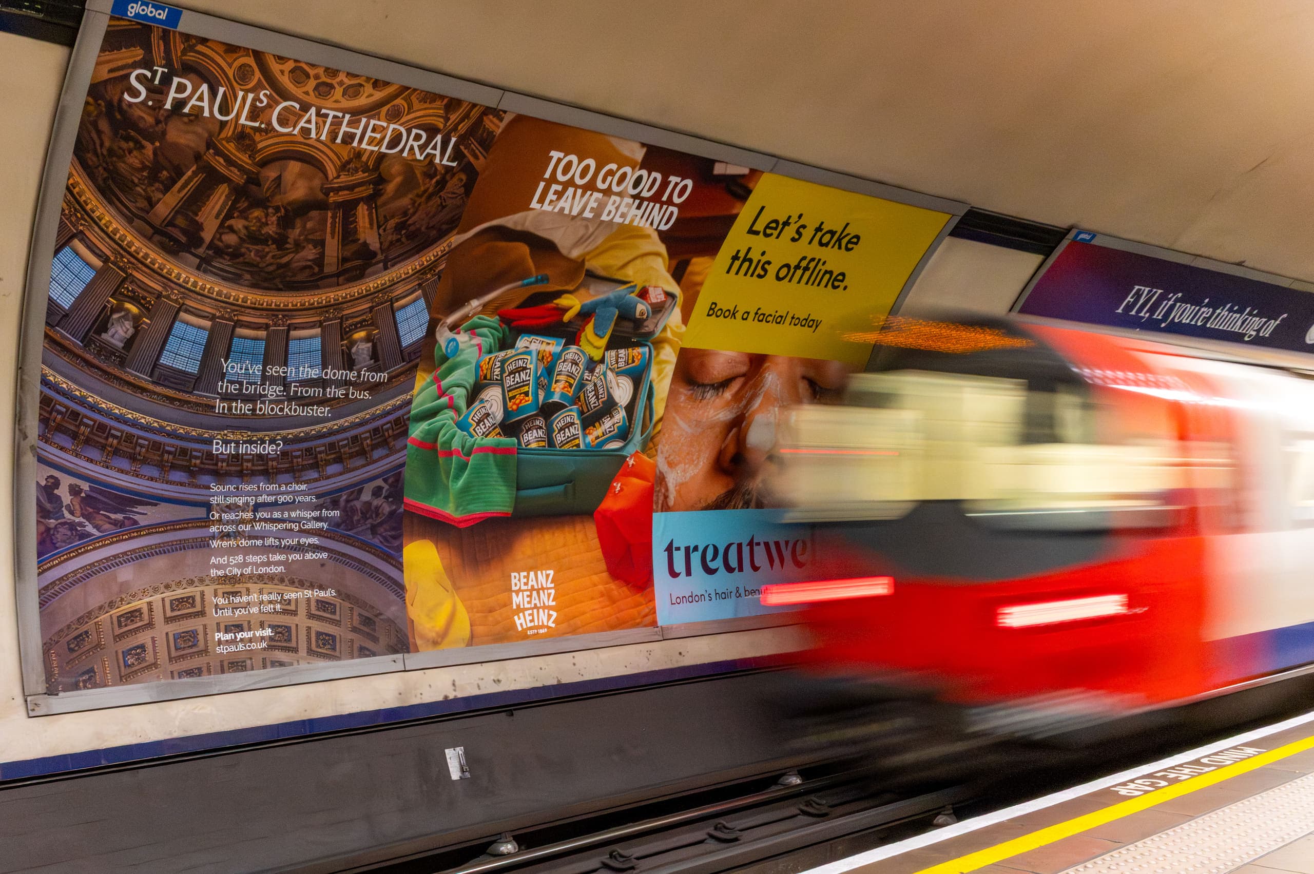 A run of cross-track poster sites along an Underground platform wall carrying several different advertisements, with a train passing at speed.