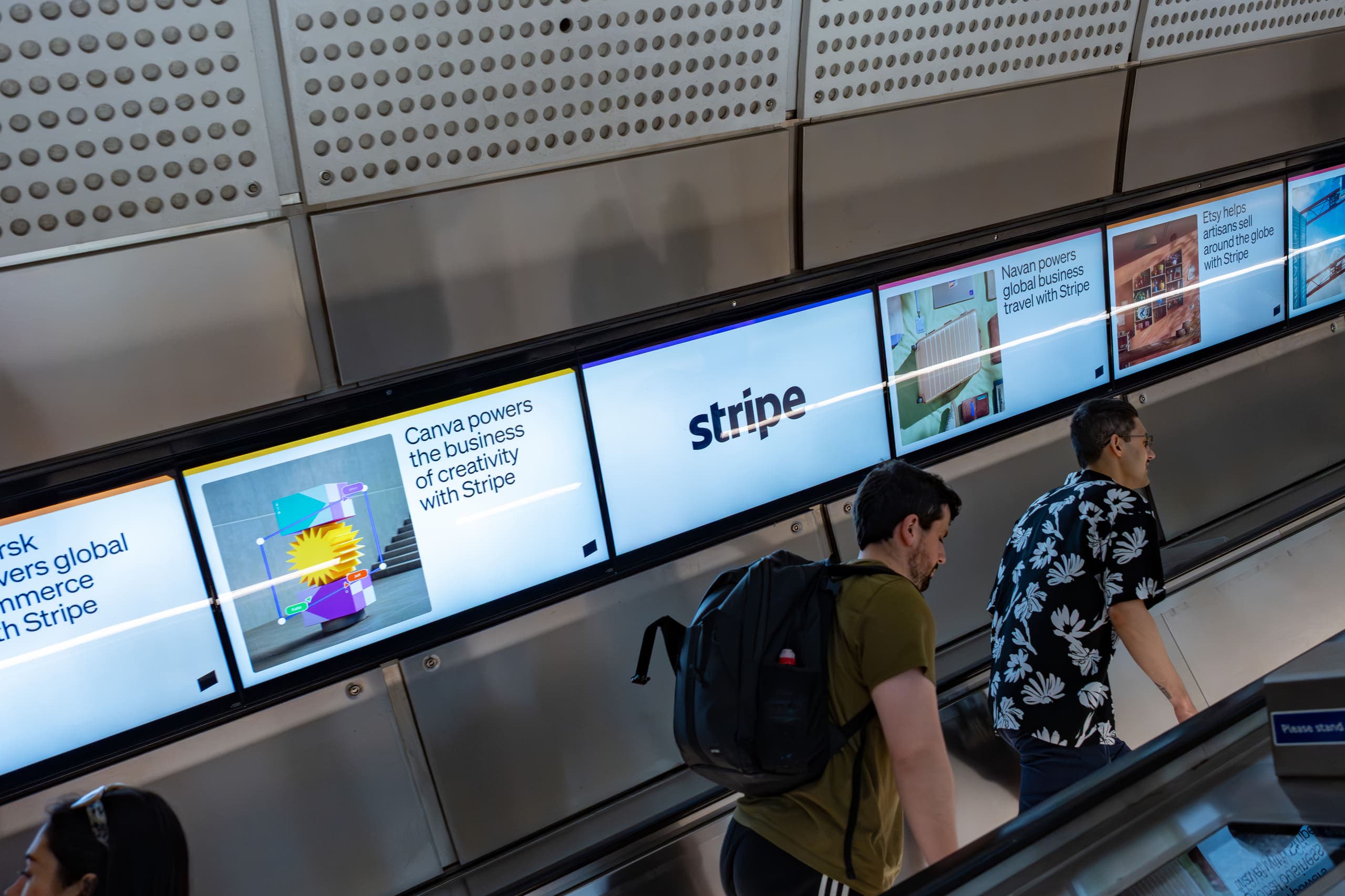A continuous run of landscape digital screens beside an Elizabeth line escalator, all carrying Stripe advertisements, as two passengers ride down.