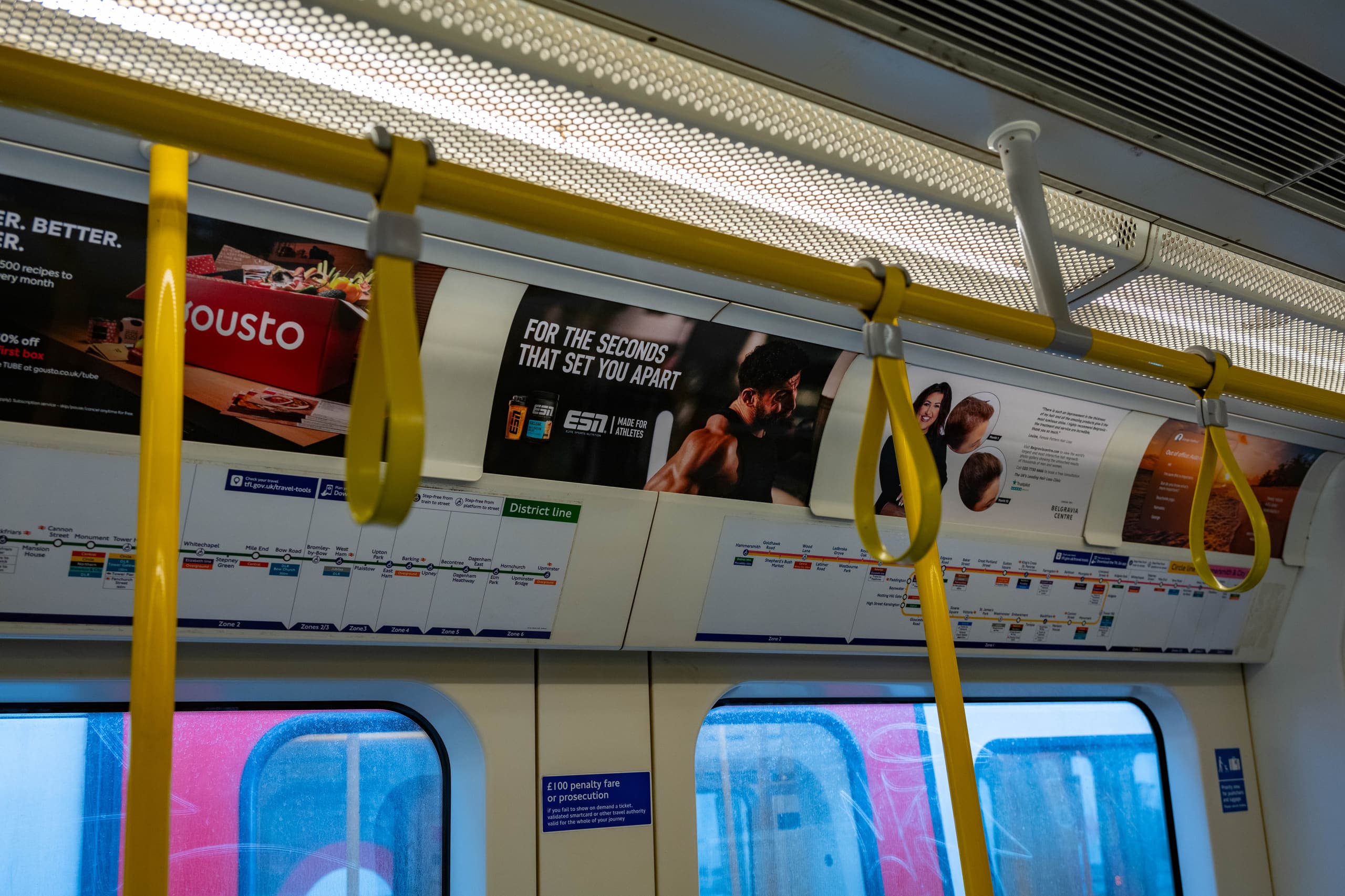 Tube car panels above the windows of a District line carriage, one of them carrying an ESN advertisement, with yellow grab handles in the foreground.