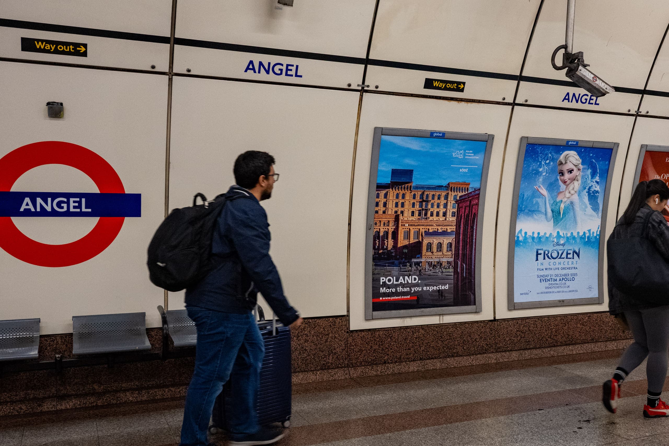 A poster site carrying a Polish Tourism Organisation advertisement on the platform wall at Angel, beside the station roundel, as a passenger passes with luggage.