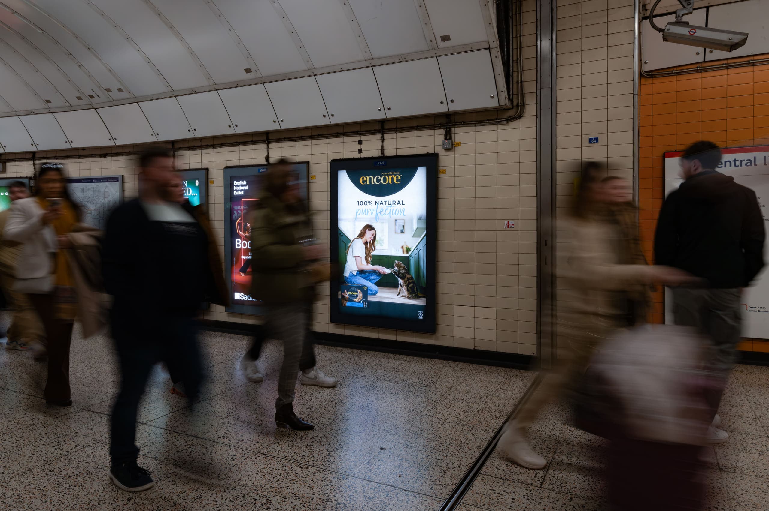 A backlit 6-sheet poster advertising Encore in a Central line passageway at Bond Street, with commuters passing in front of it in motion blur.