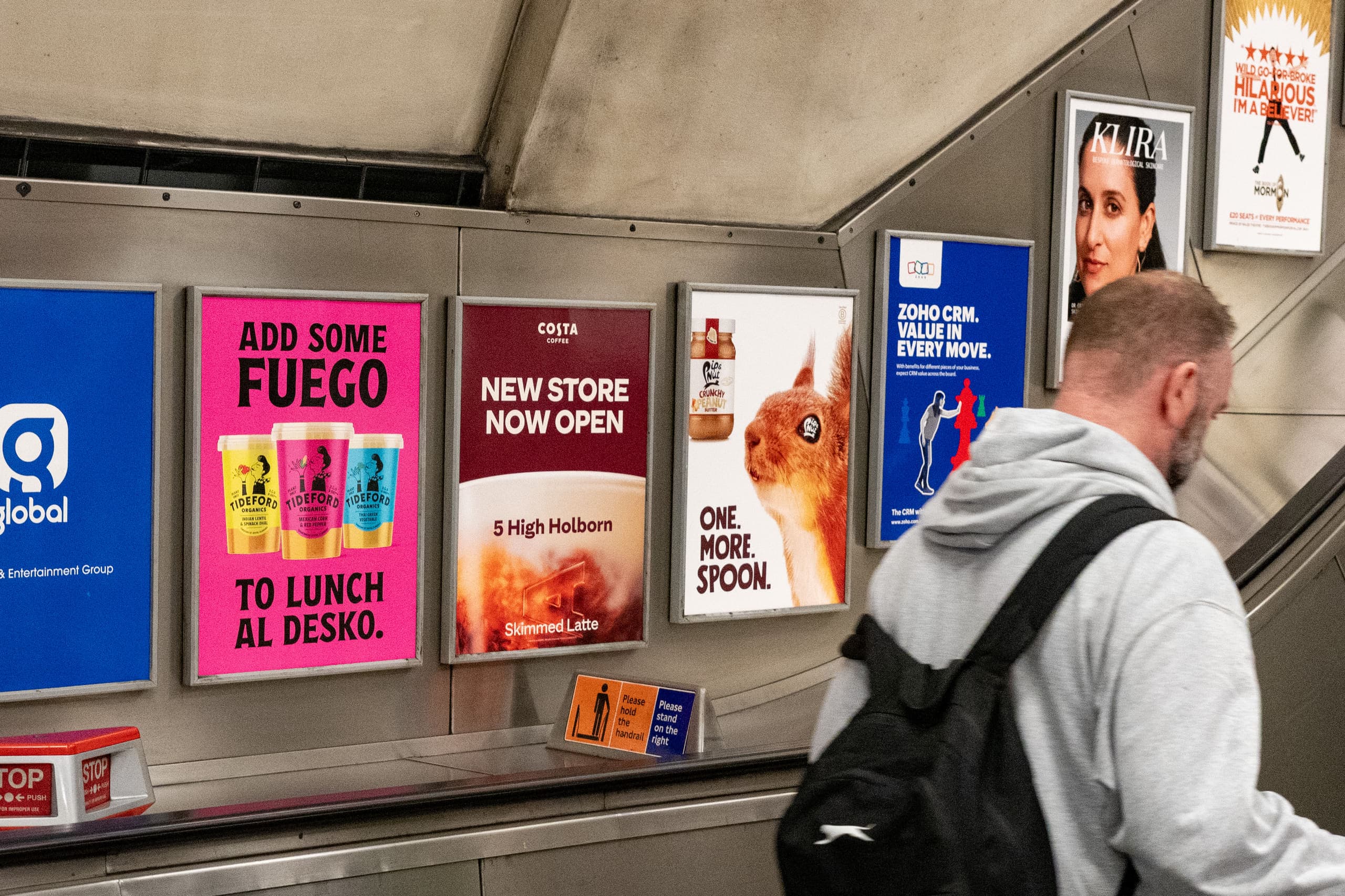 A run of static escalator panels beside an Underground escalator carrying several different advertisements, including one for Costa Coffee, as a passenger travels down.