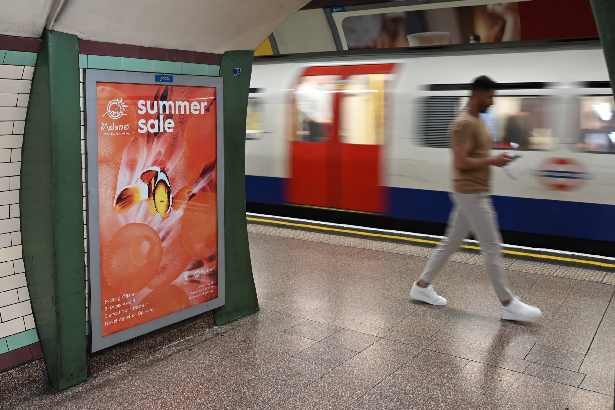 A framed portrait Visit Maldives summer sale poster on a London Underground platform wall between green pillars, with a train at the platform.