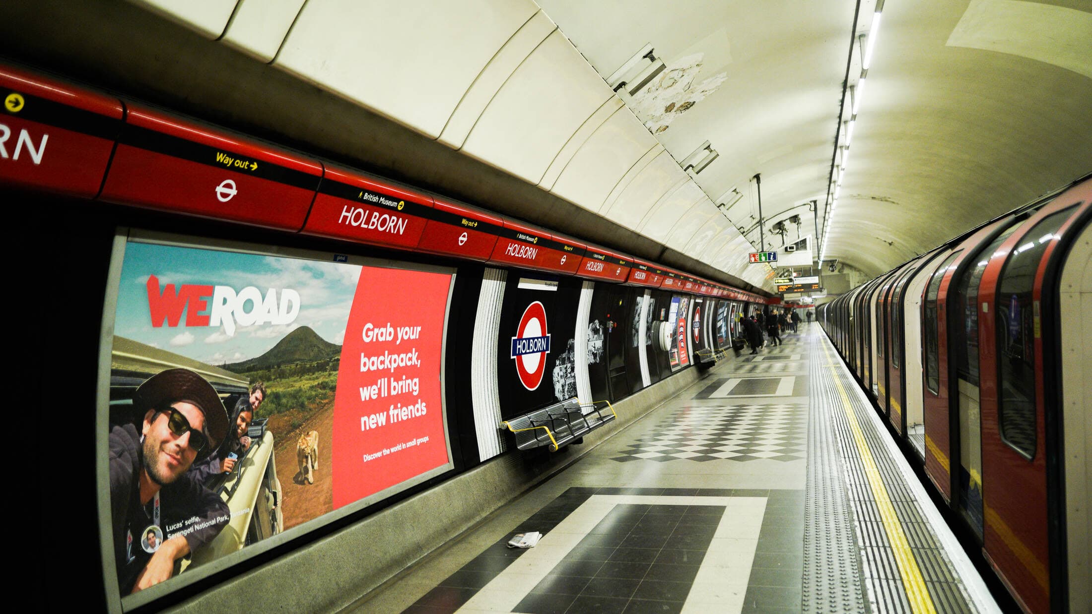 A landscape 12-sheet poster on the platform wall at Holborn carrying a WeRoad advertisement, with the station name frieze running above it and a train at the platform.