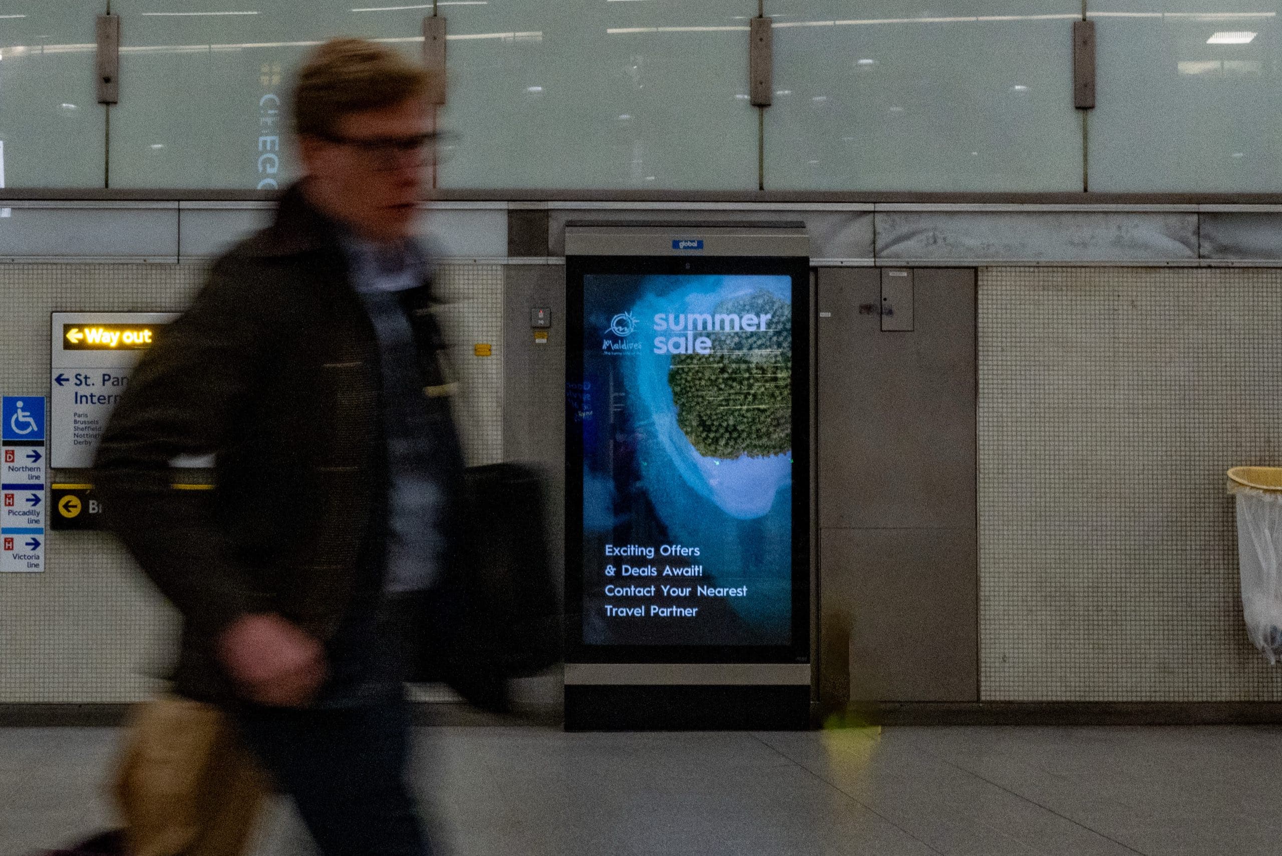 A portrait digital screen carrying a Visit Maldives summer sale advertisement in a King’s Cross St Pancras concourse, with a commuter walking past.