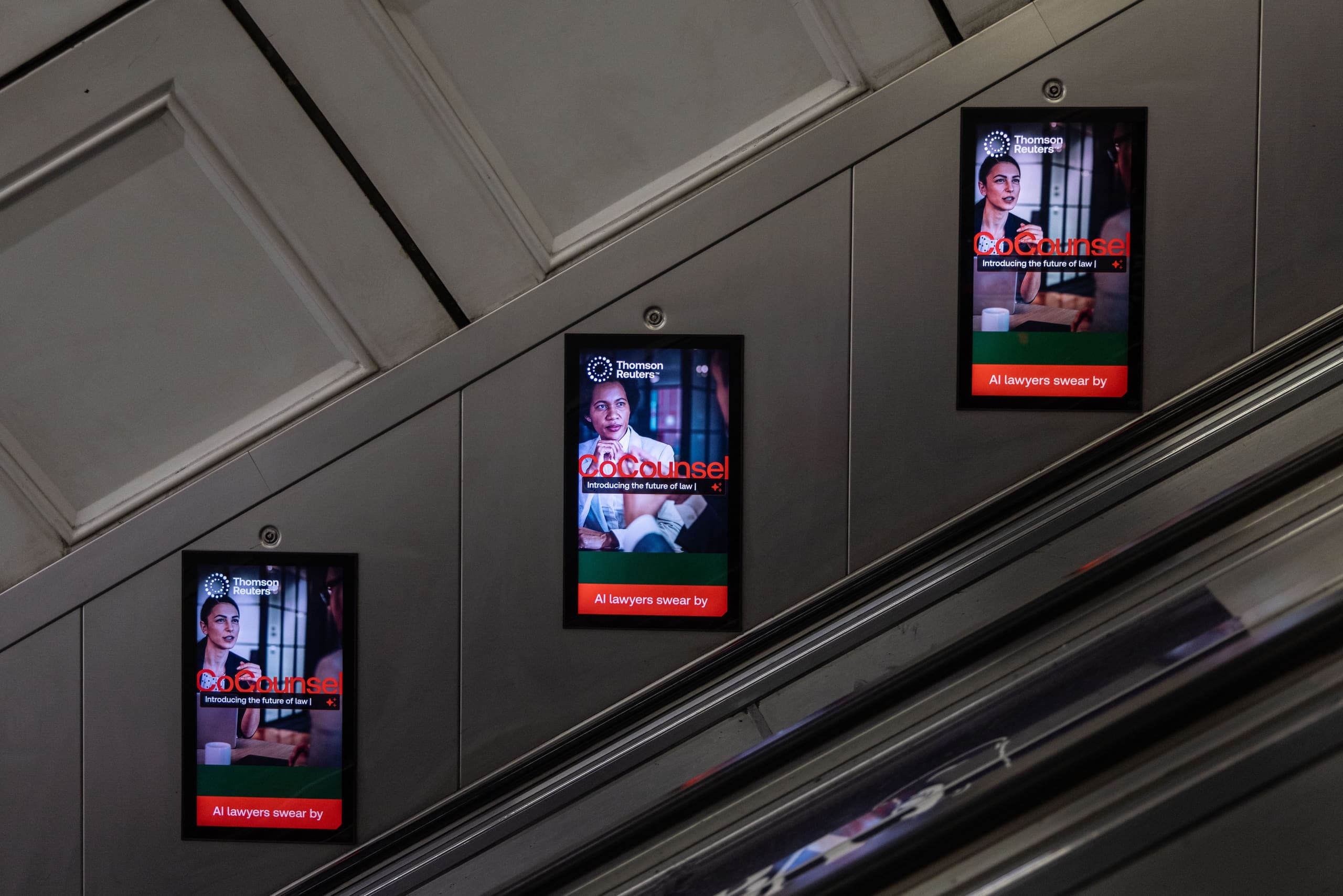 Three digital escalator panels in sequence on the wall beside an Underground escalator, each carrying a Thomson Reuters advertisement.