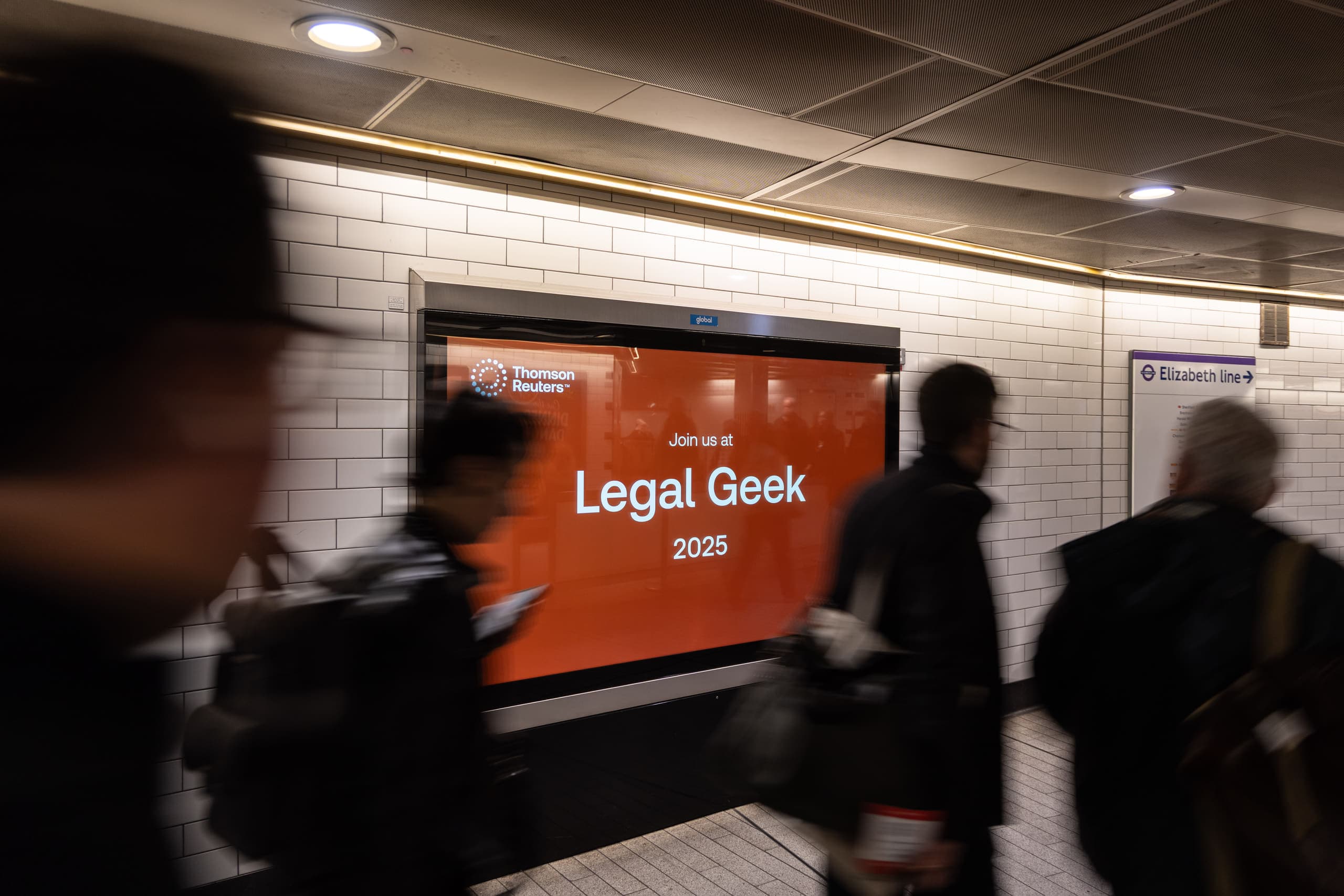A large-format digital screen carrying a Thomson Reuters advertisement on a tiled wall at Liverpool Street, beside an Elizabeth line direction sign, as commuters pass.