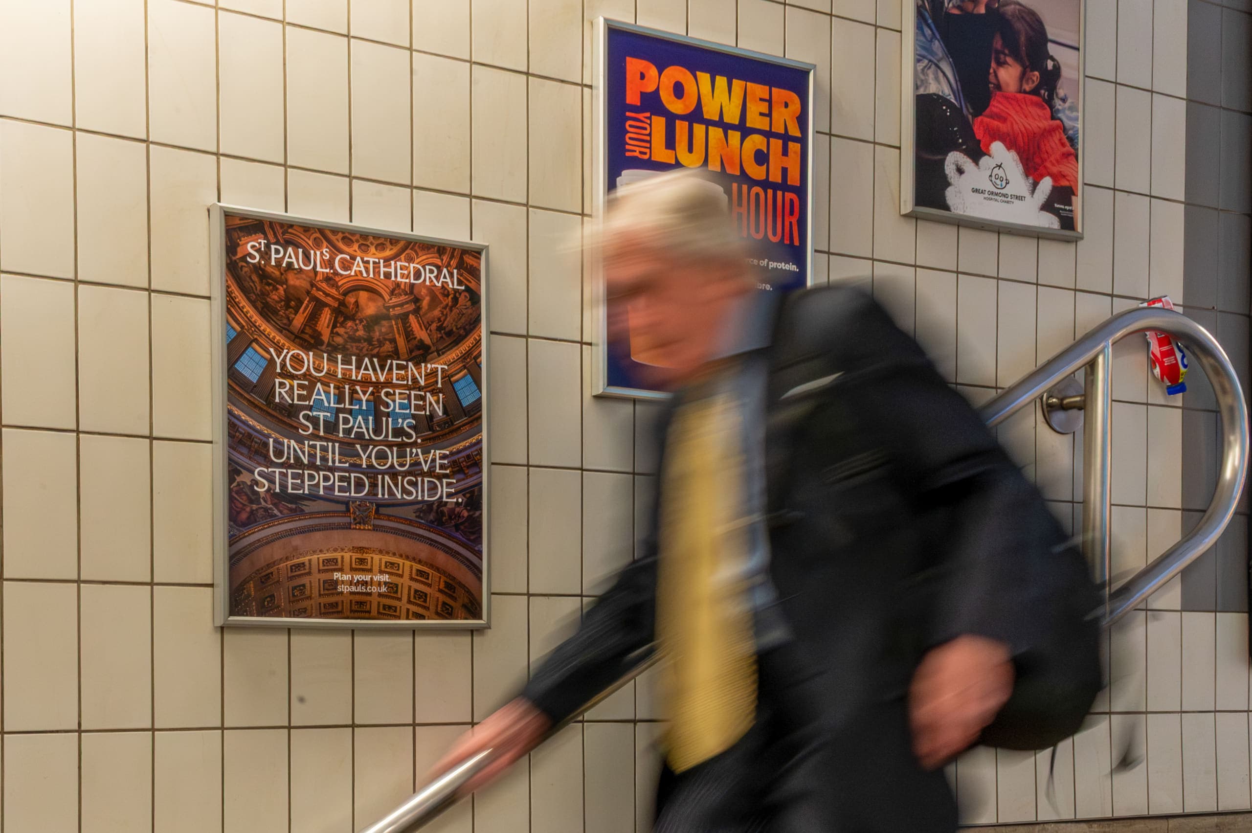 A static escalator panel advertising St Paul's Cathedral on a tiled wall, as a passenger passes in motion blur.