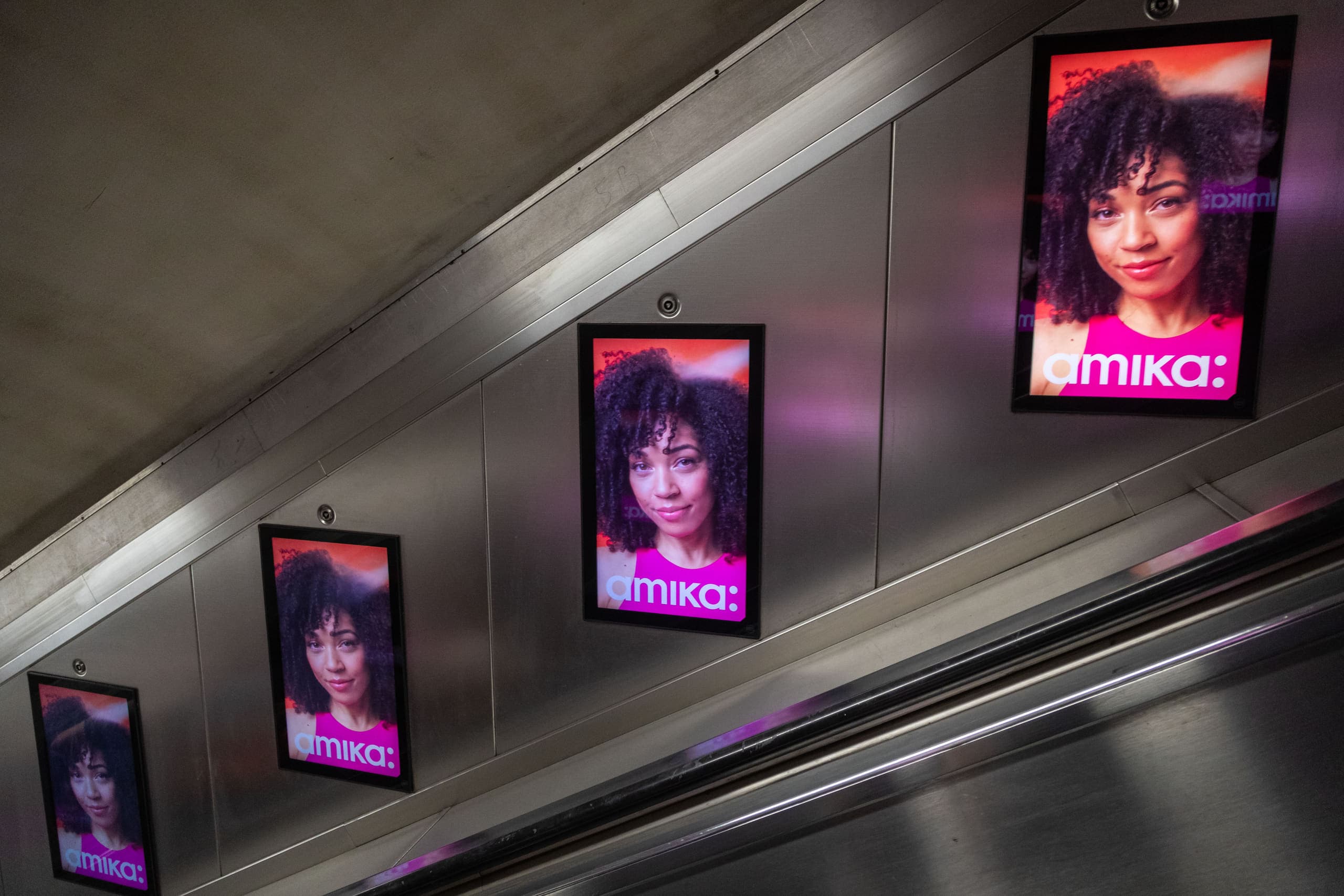 Five digital escalator panels running up the wall beside an Underground escalator, each carrying the same amika advertisement.