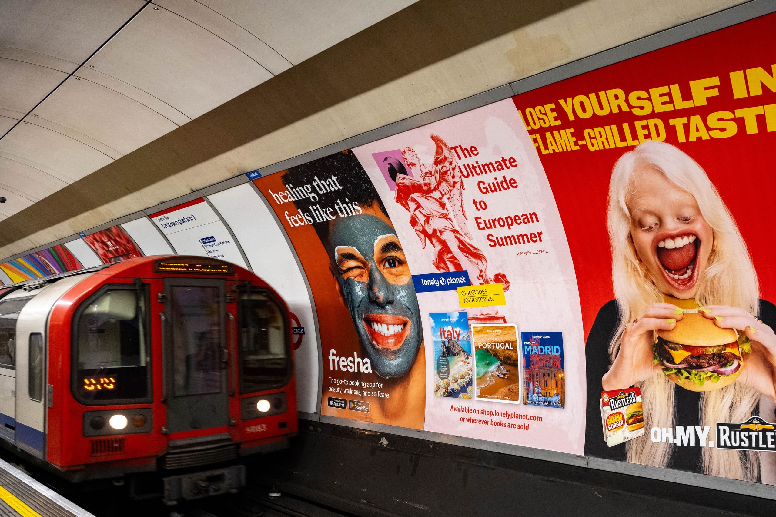 A run of cross-track poster sites along an Underground platform wall carrying four different advertisements, with a train entering the station.