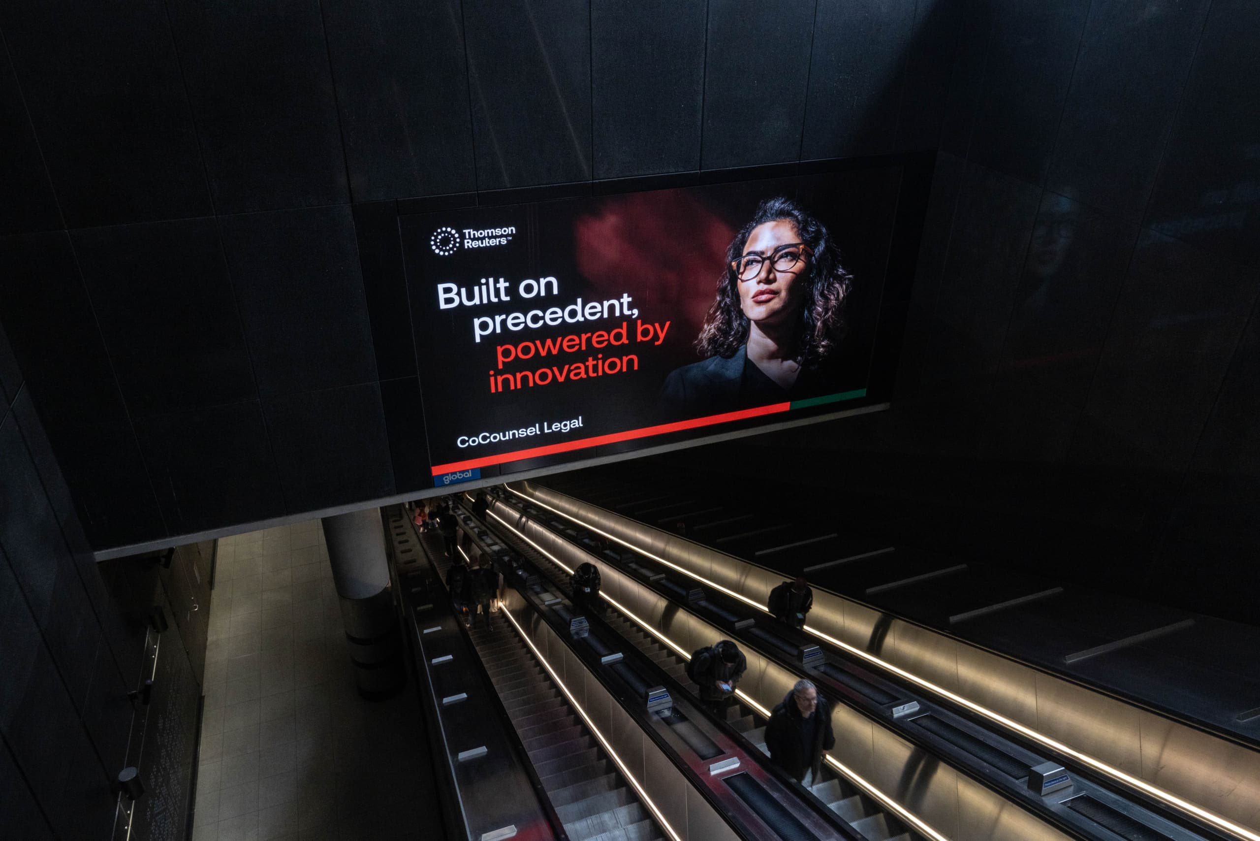 A large landscape digital screen carrying a Thomson Reuters advertisement, mounted above a bank of Underground escalators.