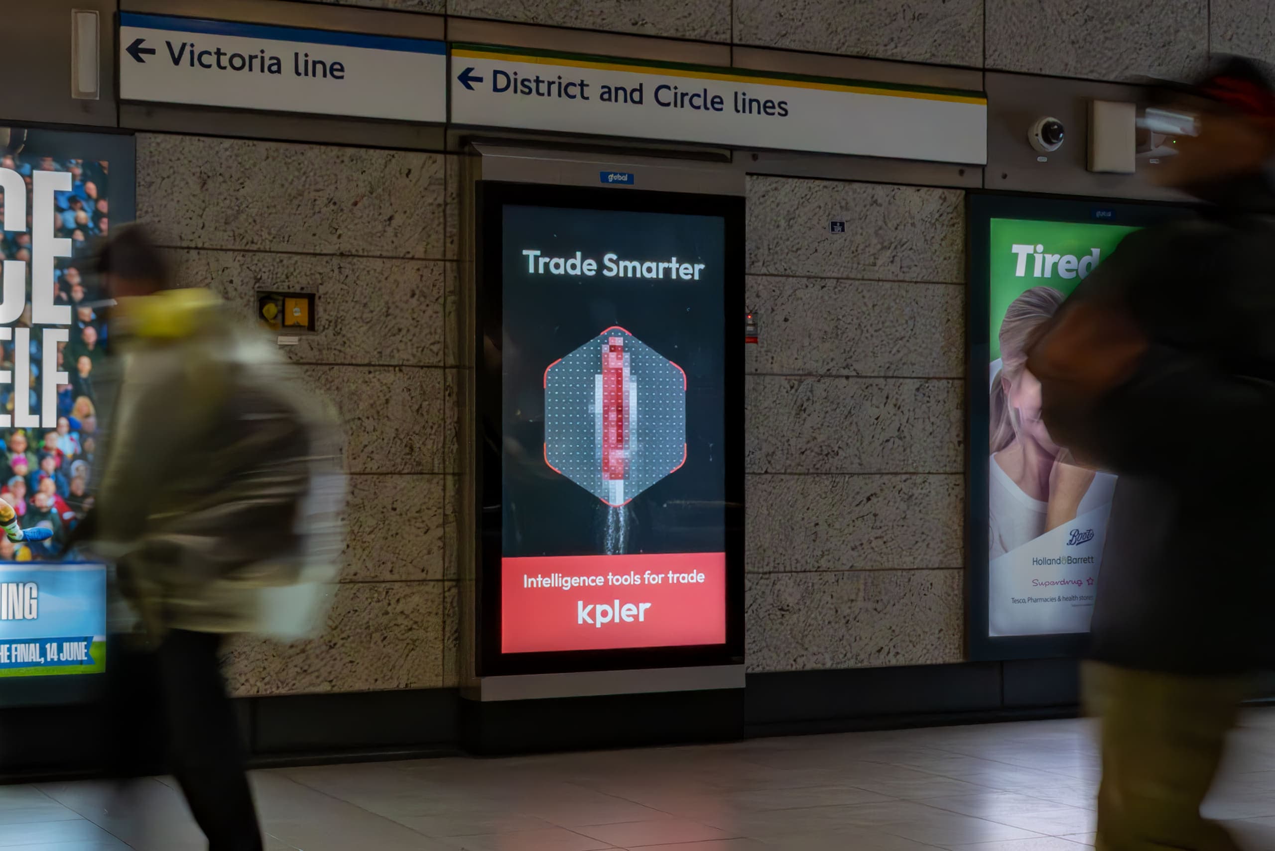 A free-standing digital 6-sheet screen carrying a Kpler advertisement at Victoria, below signs for the Victoria, District and Circle lines, with passengers crossing in motion blur.