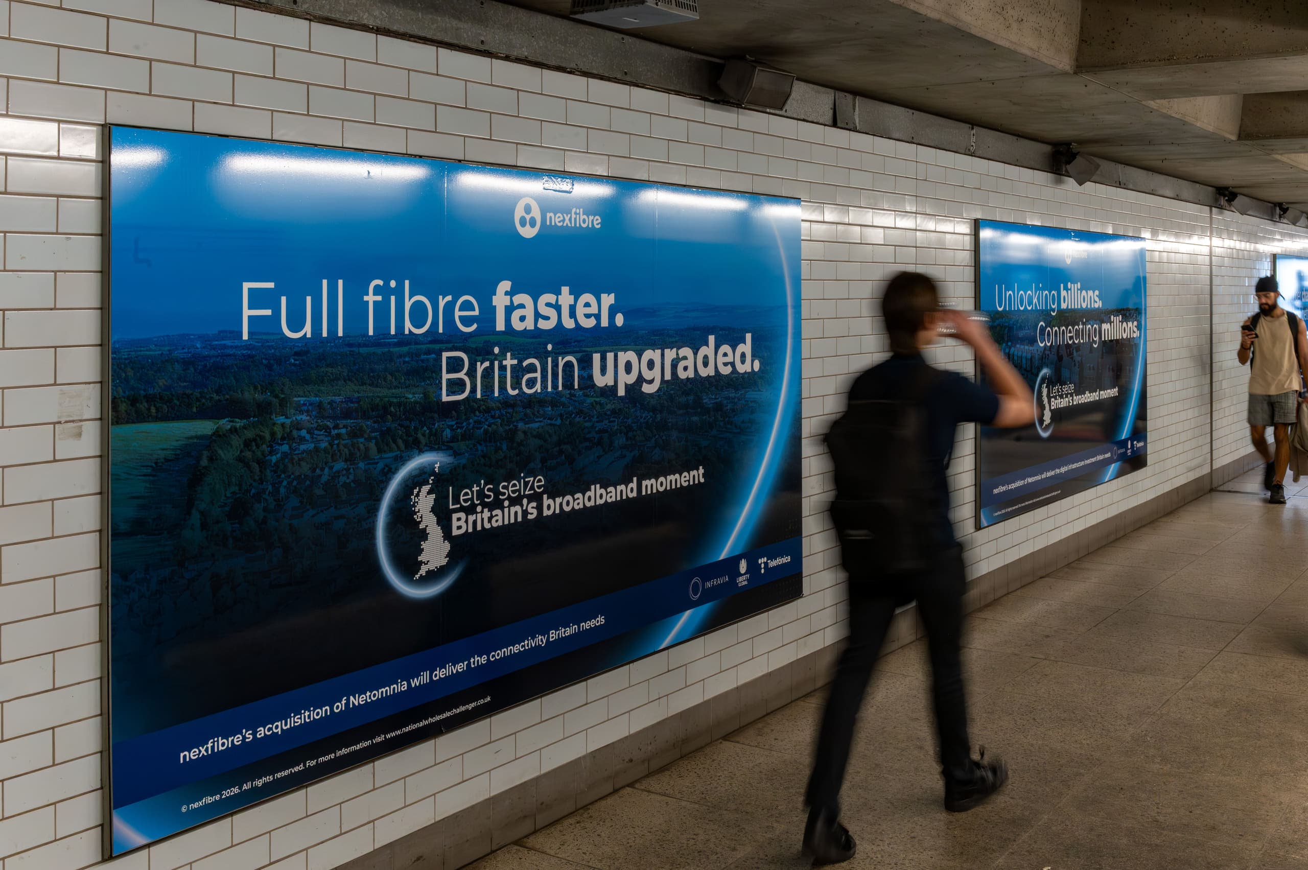 A single large-format nexfibre poster panel on the tiled wall of an Underground walkway, with a passenger walking past it.