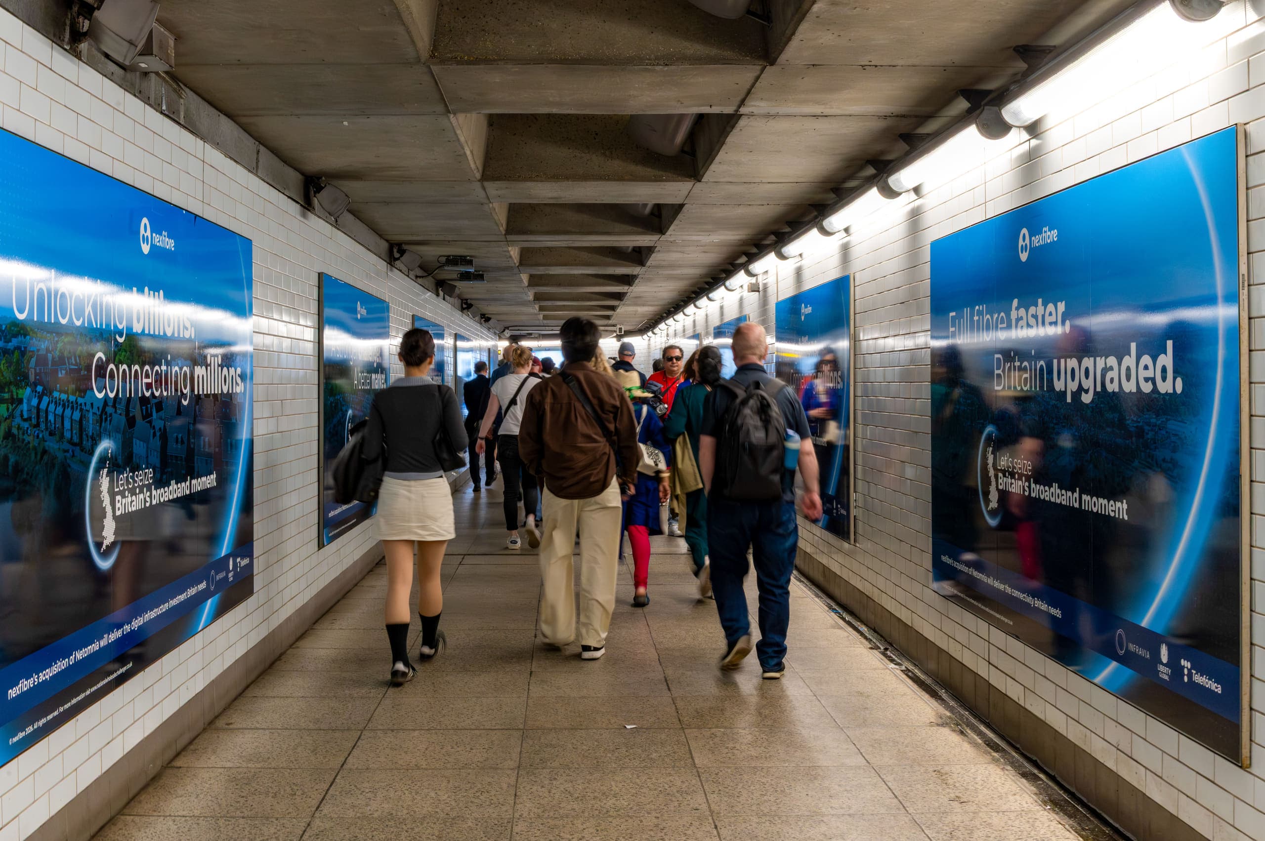 Advertising panels running the length of both walls of a tiled Underground interchange corridor, all carrying the same nexfibre campaign, as commuters walk away from the camera.