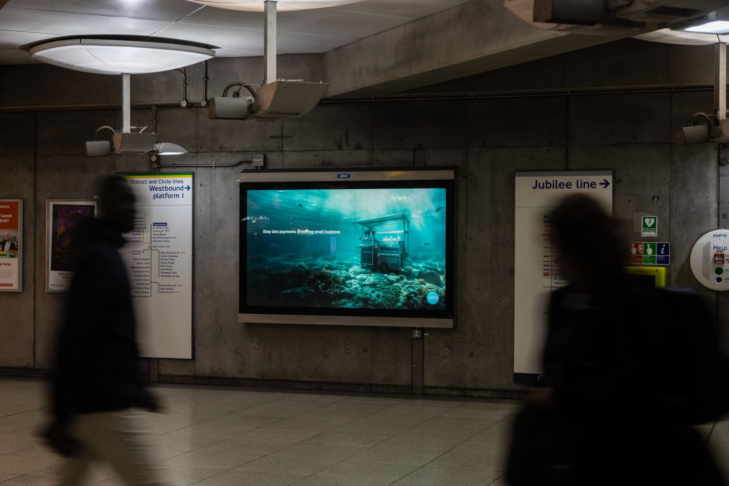A large landscape digital screen carrying a Xero advertisement against the board-marked concrete at Westminster, beside signs for the westbound District and Circle line platform and the Jubilee line.